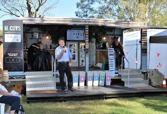 Man with microphone in front of mobile barber shop trailer with customers in chairs getting haircut