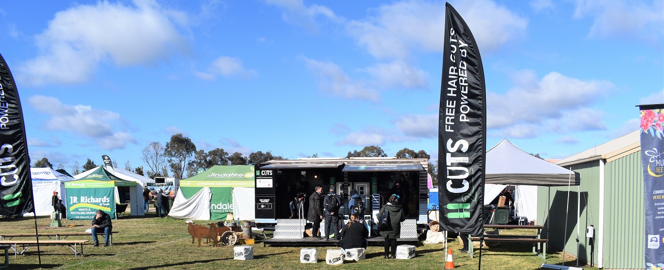 people surrounding mobile barber trailer at field day on green paddock with blue sky