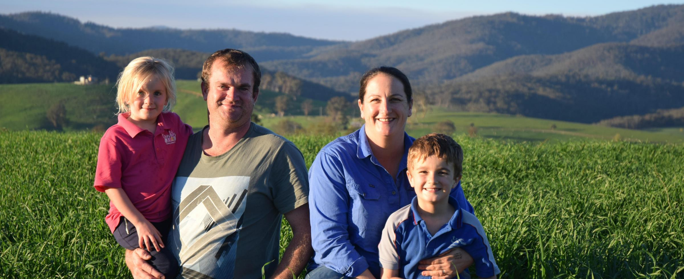 Jen and Brad Smith stand in a crop paddock on their farm. Jen is holding their son and Brad is holding their daughter.
